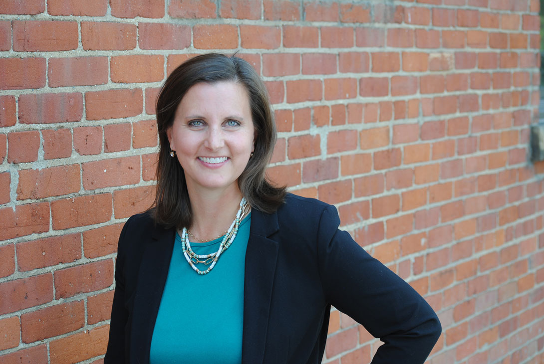 Sarah Morgan stands in front of a brick wall smiling at the camera.